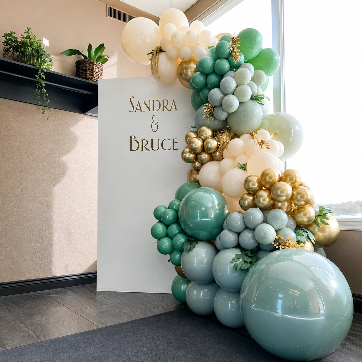 Sandra and Bruce celebration backdrop featuring sage green, white, and gold balloon garlands with greenery and elegant gold accents.