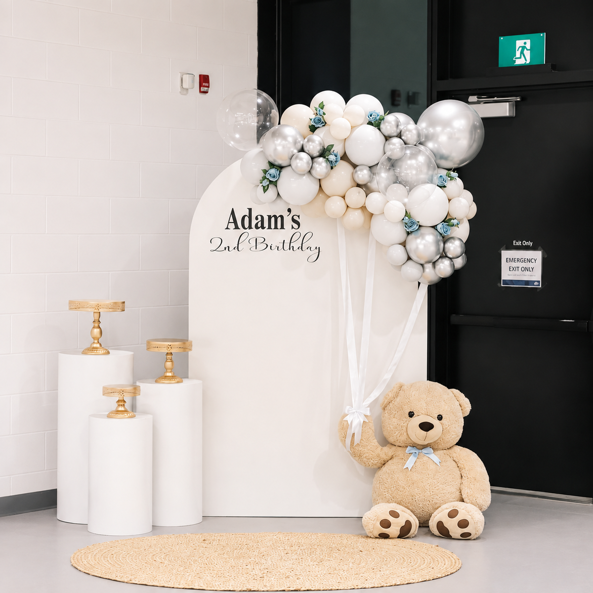 Adam’s second birthday backdrop featuring white and silver balloon decor, blue floral accents, a personalised white arch, gold dessert stands, and a large teddy bear.