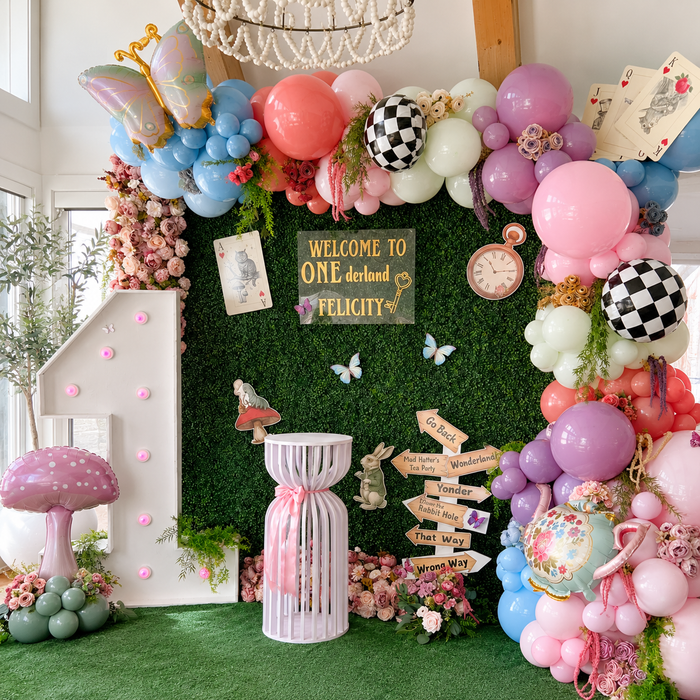 Alice in Wonderland first birthday backdrop featuring pastel balloon garlands, playing cards, butterflies, flowers, and a large illuminated number one.