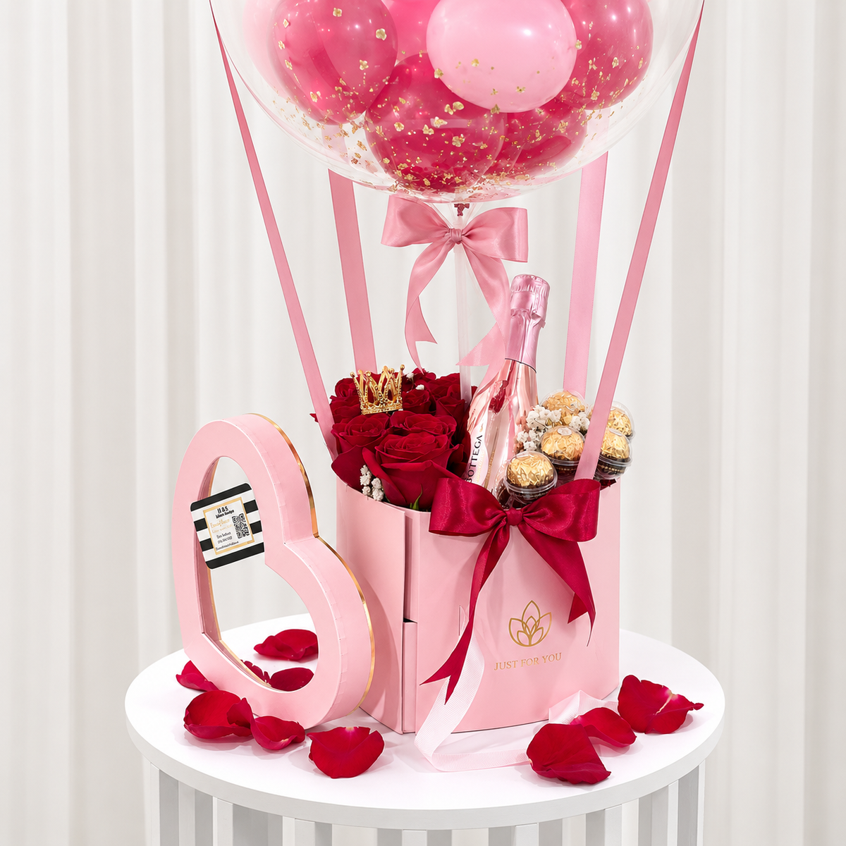 Close-up of a romantic anniversary gift featuring red roses, chocolates, a pink sparkling beverage bottle, heart-shaped decor, pink and burgundy ribbons, and a balloon arrangement.