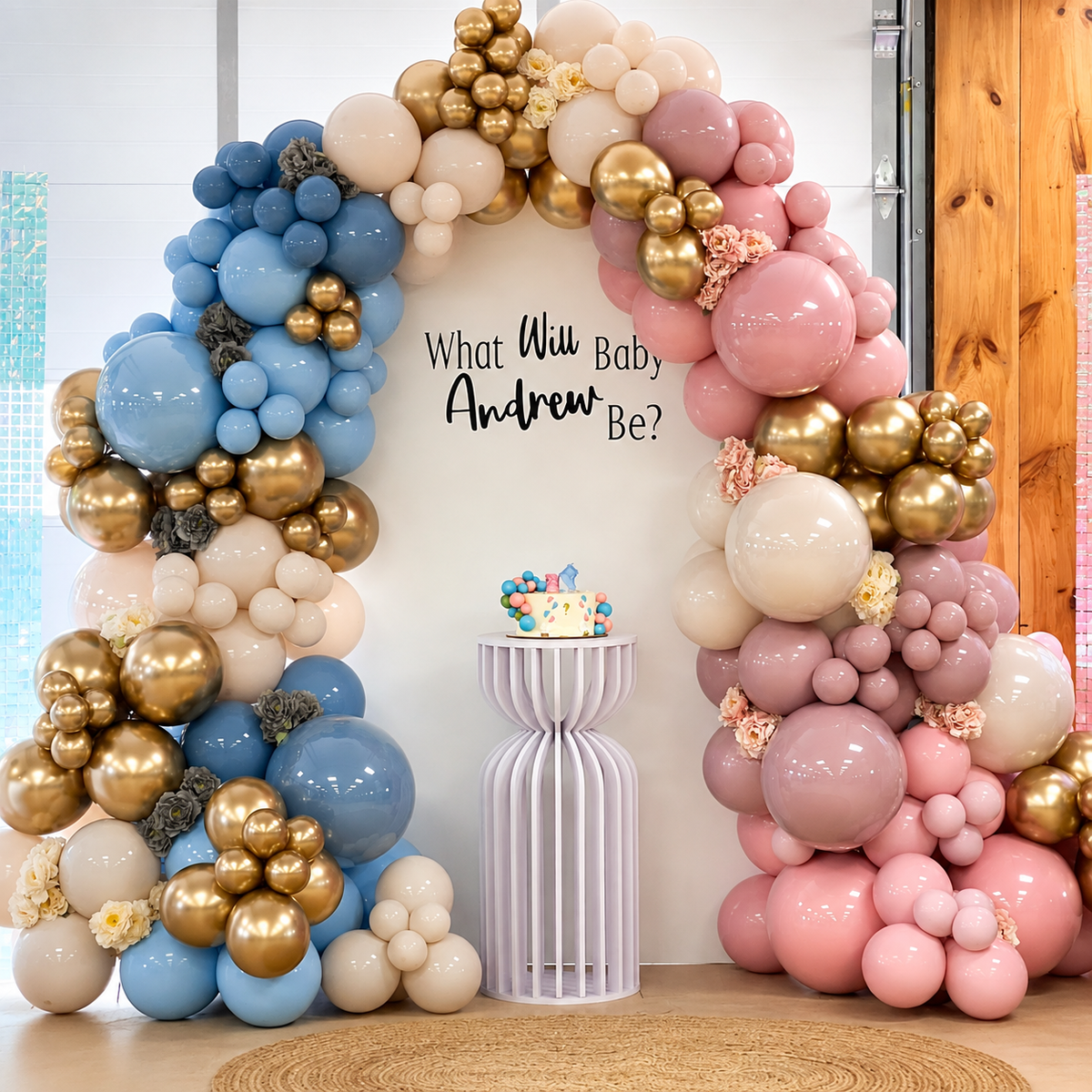 Baby Andrew gender reveal backdrop featuring blue, pink, cream, and gold balloon garlands, floral accents, a white backdrop, and a central reveal cake pedestal.
