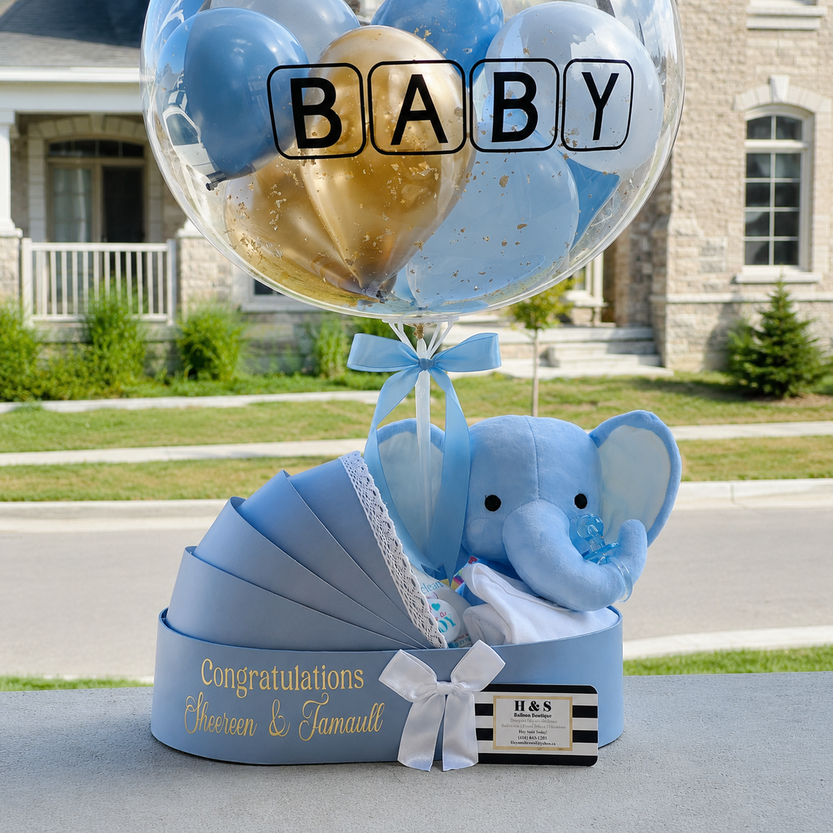 Baby boy gift arrangement featuring a personalised clear balloon with blue, white, and gold balloons, a plush elephant, baby essentials, and a blue pram-shaped gift box.