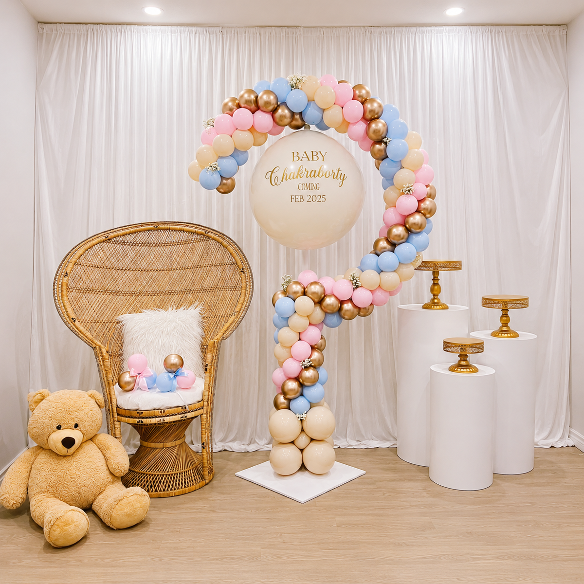 Chakraborty shower backdrop featuring a pastel pink, blue, cream, and gold balloon arch, personalized “Coming Feb 2025” balloon, rattan peacock chair, teddy bear, and white pedestal stands.