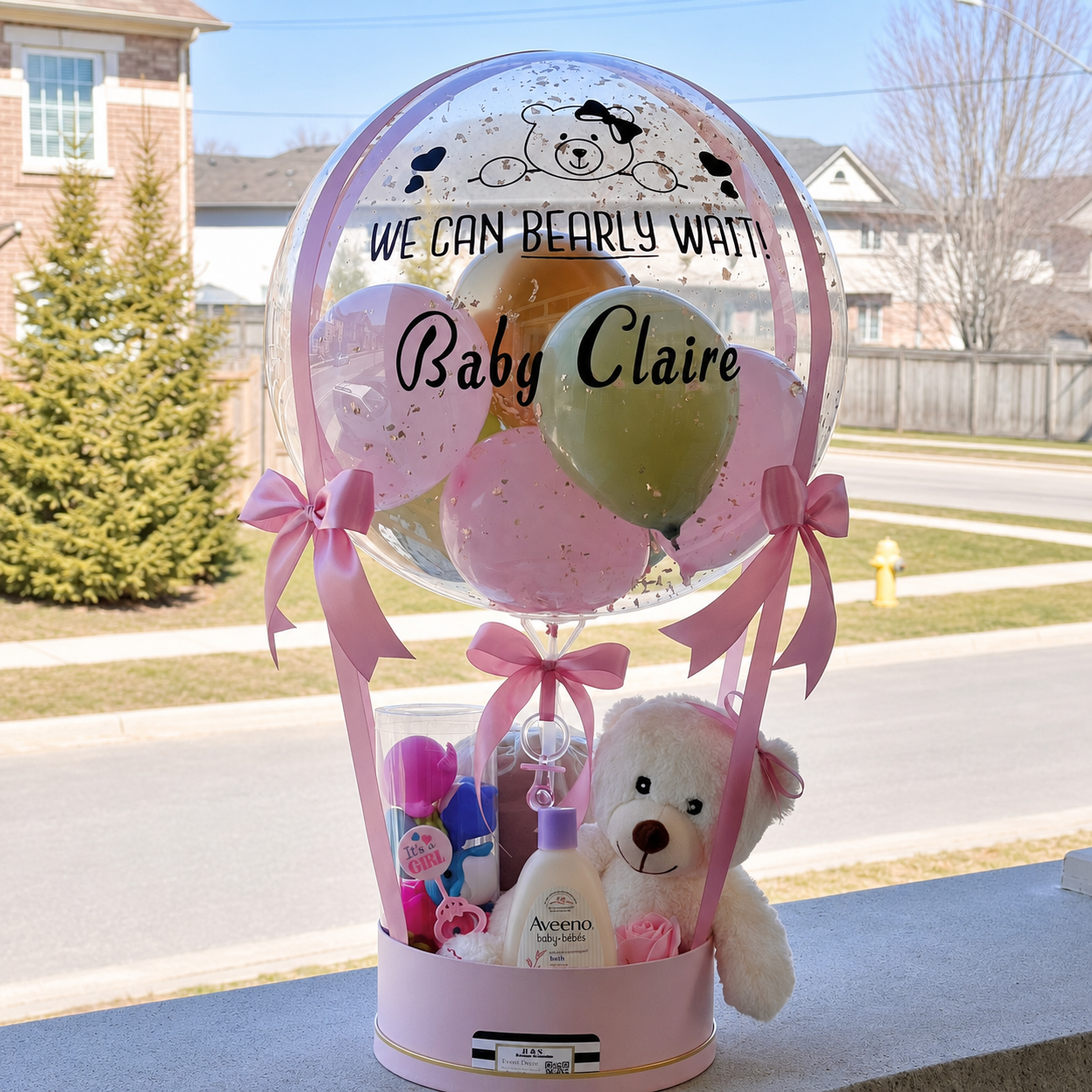 Baby girl gift arrangement for Claire featuring a personalised clear balloon, pastel balloons, pink ribbons, a teddy bear, baby bath products, and newborn gifts.