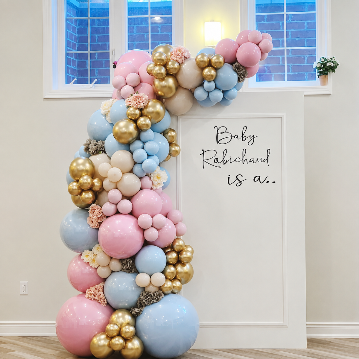 Baby gender reveal backdrop featuring pink, blue, cream, and gold balloon garlands with floral accents and a personalised “Baby Rabichaud is a...” sign.