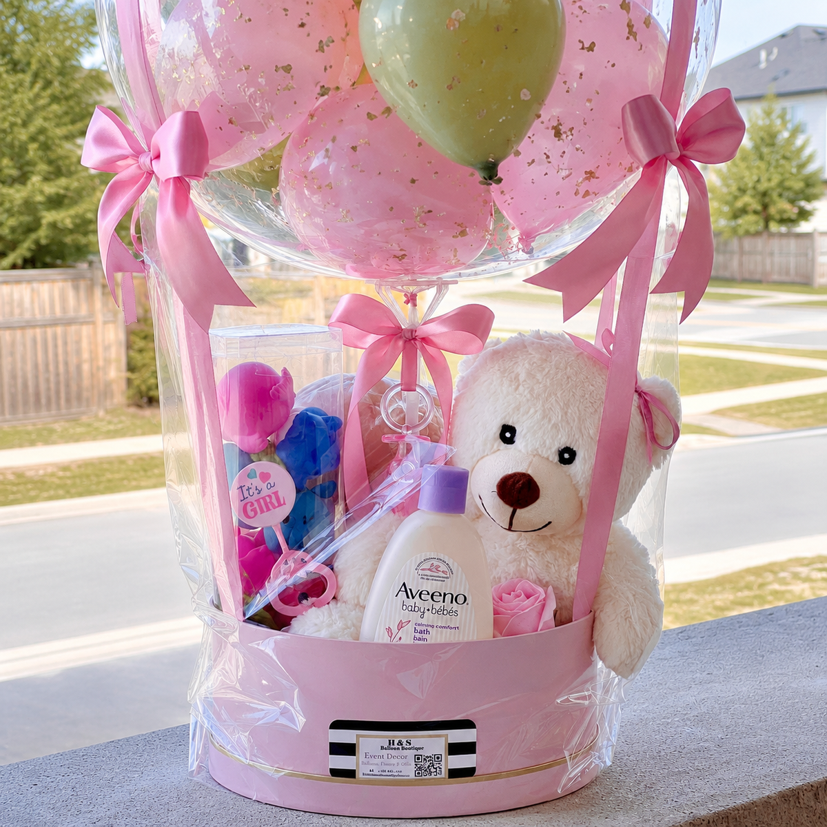 Close-up of a baby girl gift basket featuring a teddy bear, baby bath products, pink ribbons, and pastel balloons inside a clear balloon arrangement.