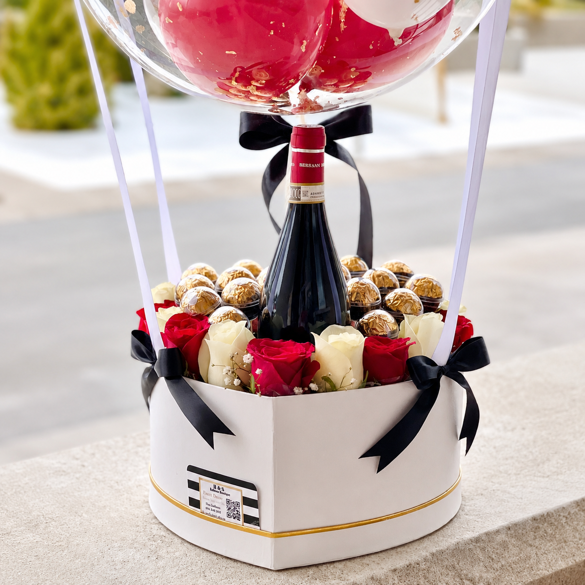 Close-up of a birthday gift arrangement featuring a wine bottle, red and white roses, chocolates, black ribbon accents, and a personalised balloon.