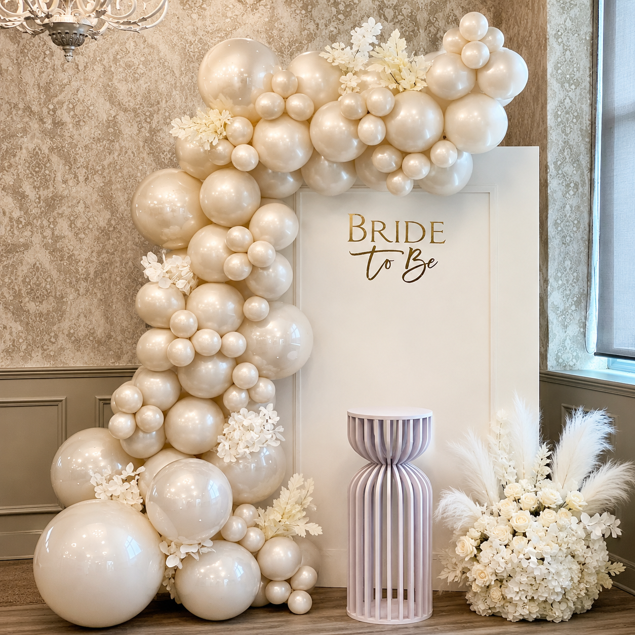 Elegant bride-to-be backdrop featuring an all-white balloon garland, white floral accents, a white panel with gold lettering, and a modern white pedestal.