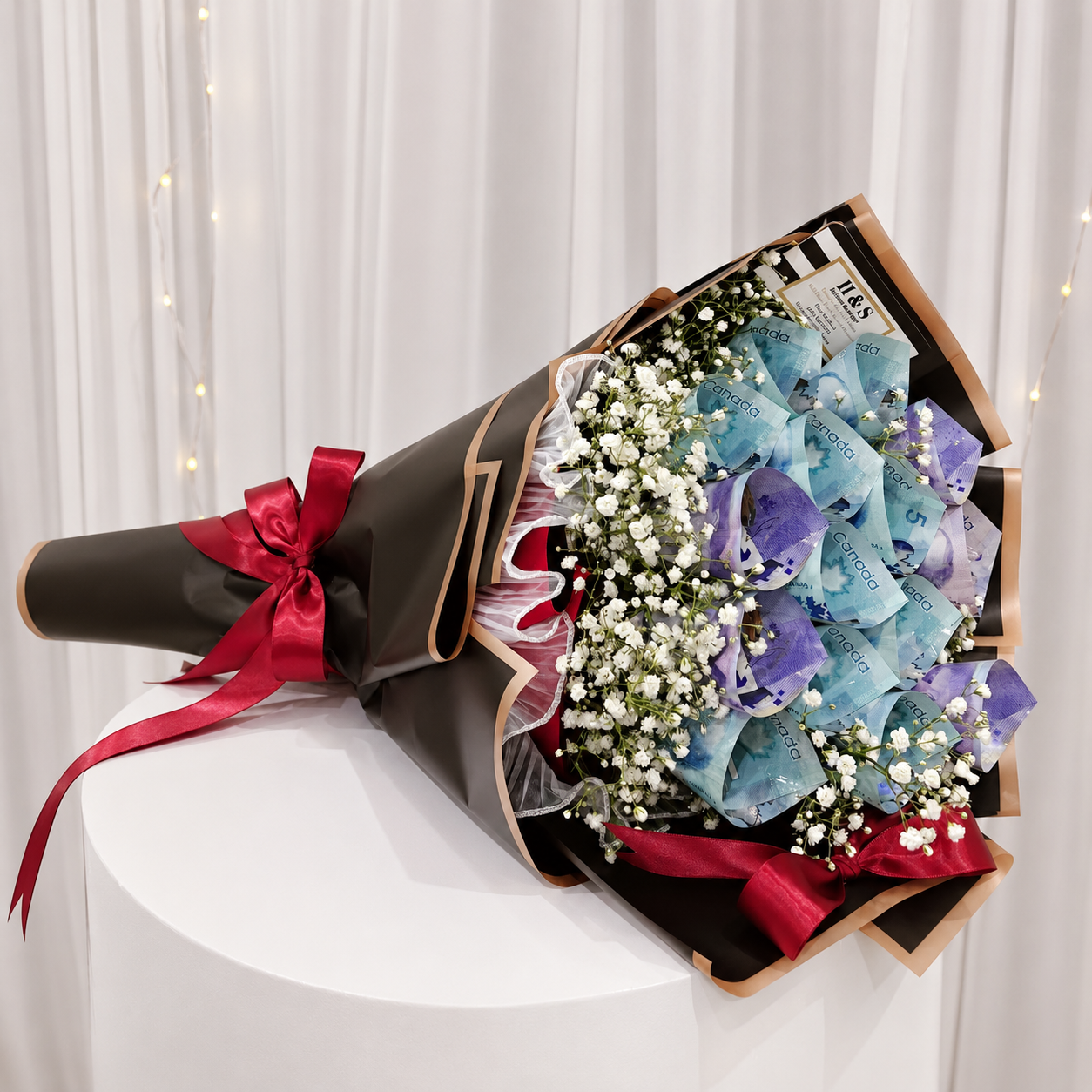 Luxury money bouquet featuring folded Canadian banknotes, white baby’s breath flowers, black wrapping with gold trim, and deep red satin ribbon bows.