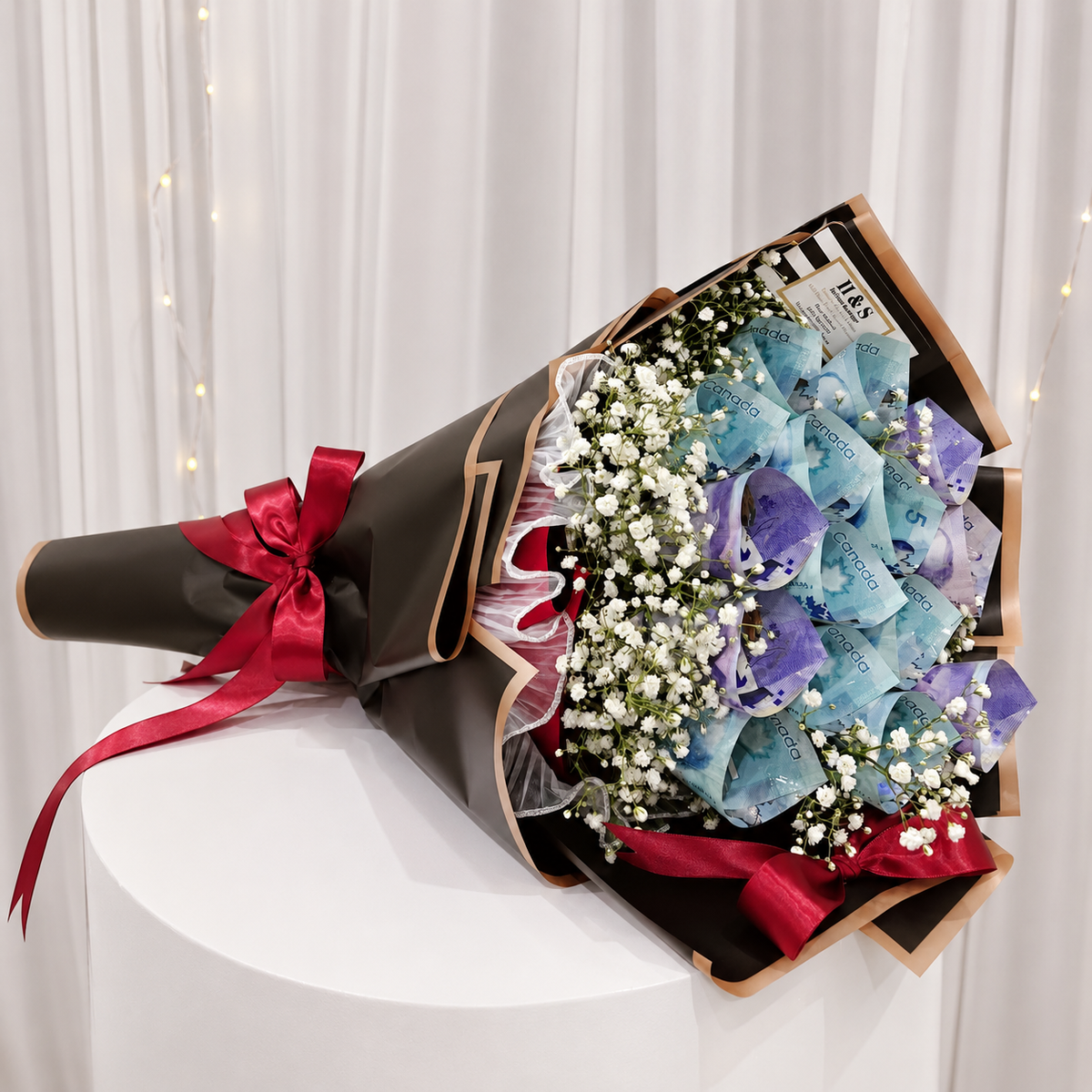 Luxury money bouquet featuring folded Canadian banknotes, white baby’s breath flowers, black wrapping with gold trim, and deep red satin ribbon bows.