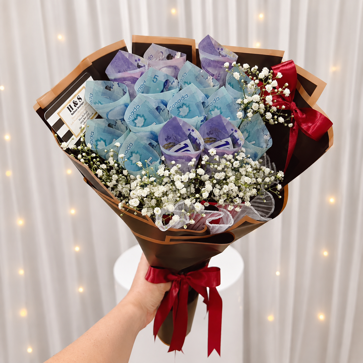 Luxury money bouquet featuring folded Canadian banknotes, white baby’s breath flowers, black wrapping with gold trim, and deep red satin ribbon bows.
