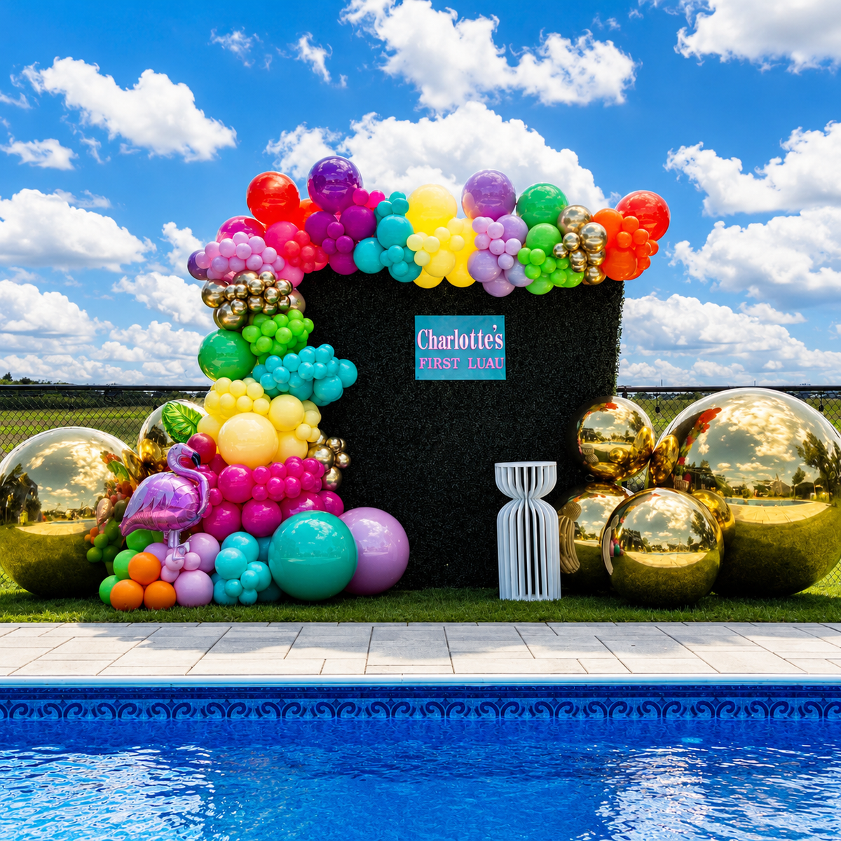 Charlotte’s first luau poolside backdrop featuring a colorful rainbow balloon garland, tropical accents, black greenery wall, and oversized gold balloons.