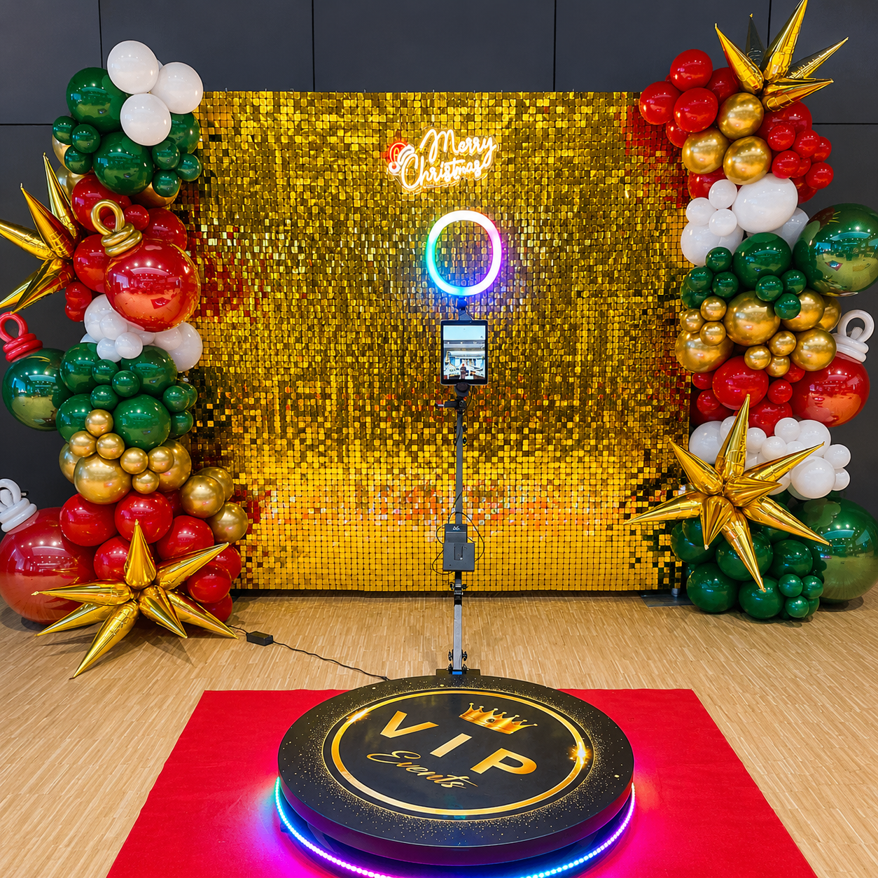 Christmas VIP 360 photo booth setup featuring a gold shimmer wall, red, green, white, and gold balloon garlands, metallic star and ornament balloons, a Merry Christmas neon sign, ring light, and branded VIP platform.