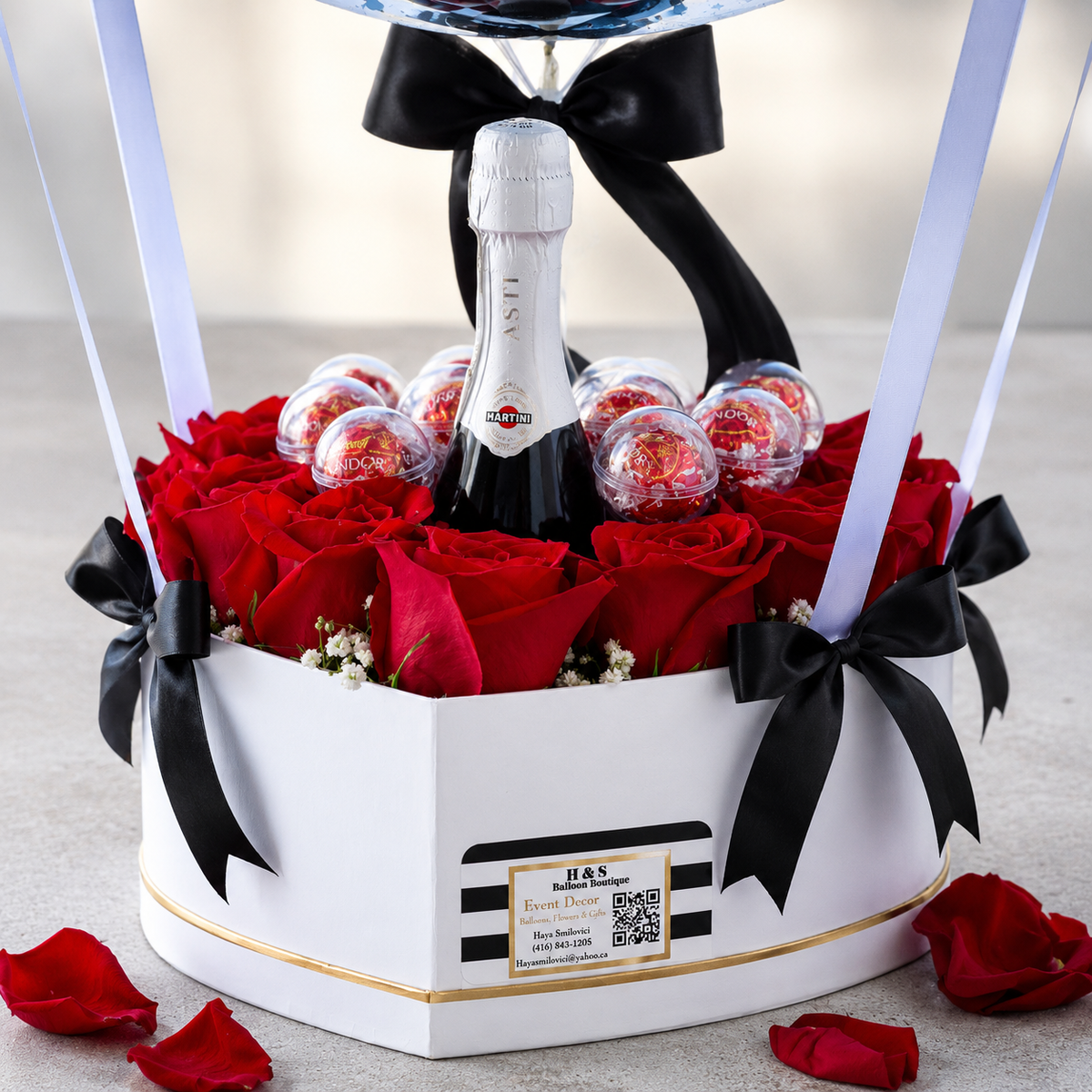 Close-up of a luxury gift arrangement featuring red roses, chocolates, a sparkling wine bottle, black satin bows, and a white heart-shaped gift box.