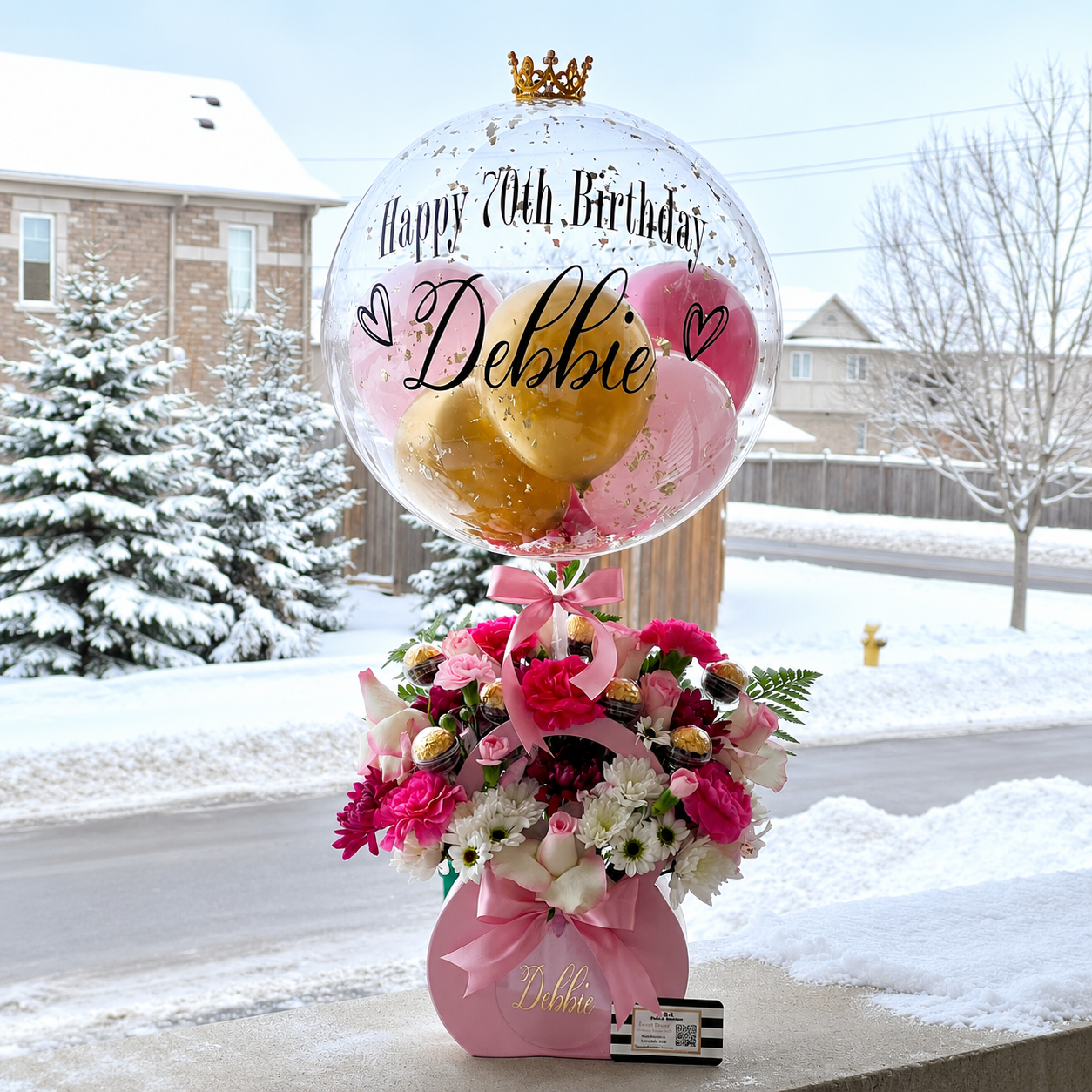 Debbie’s 70th birthday gift arrangement featuring a personalised clear balloon, pink and gold balloons, pink and white flowers, chocolates, and ribbon accents.