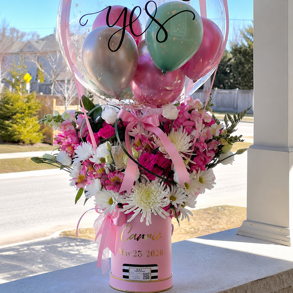 Close-up of an engagement gift arrangement featuring pink and white flowers, a personalised clear balloon with pastel balloons, and elegant pink ribbons.