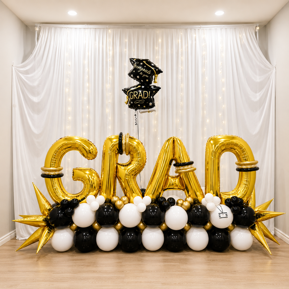 Graduation party backdrop featuring giant gold GRAD letter balloons, black, white, and gold balloon decor, star balloons, and graduation cap balloons.