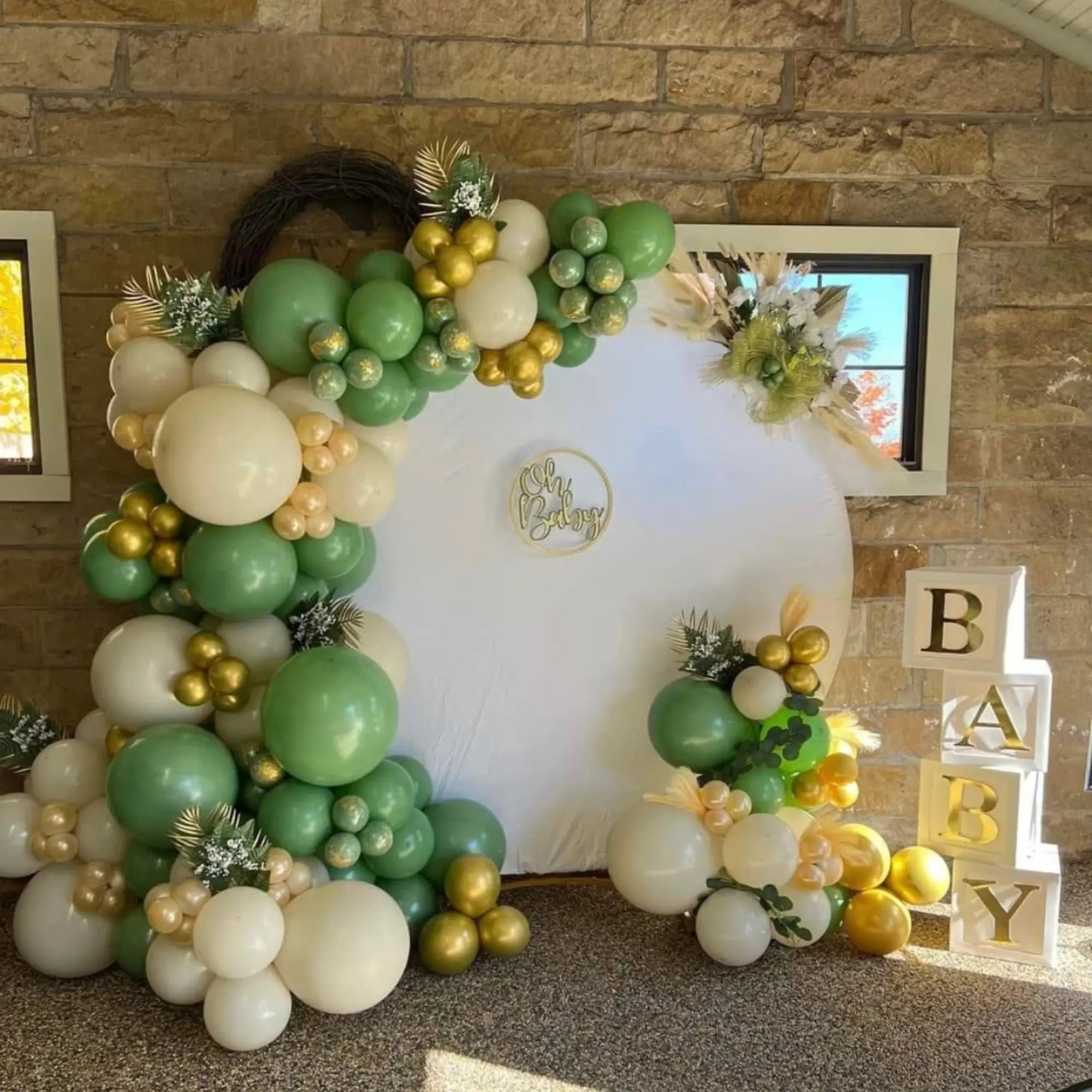 “Oh Baby” baby shower backdrop featuring sage green, white, and metallic gold balloon garlands, greenery and floral accents, a white circular backdrop, and decorative BABY blocks.