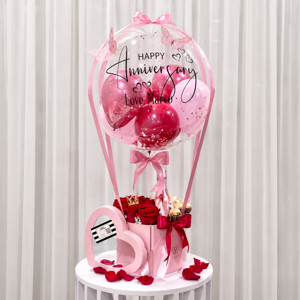 Romantic anniversary gift arrangement featuring a personalised clear balloon, pink and red balloons, red roses, chocolates, a pink bottle, and ribbon accents.