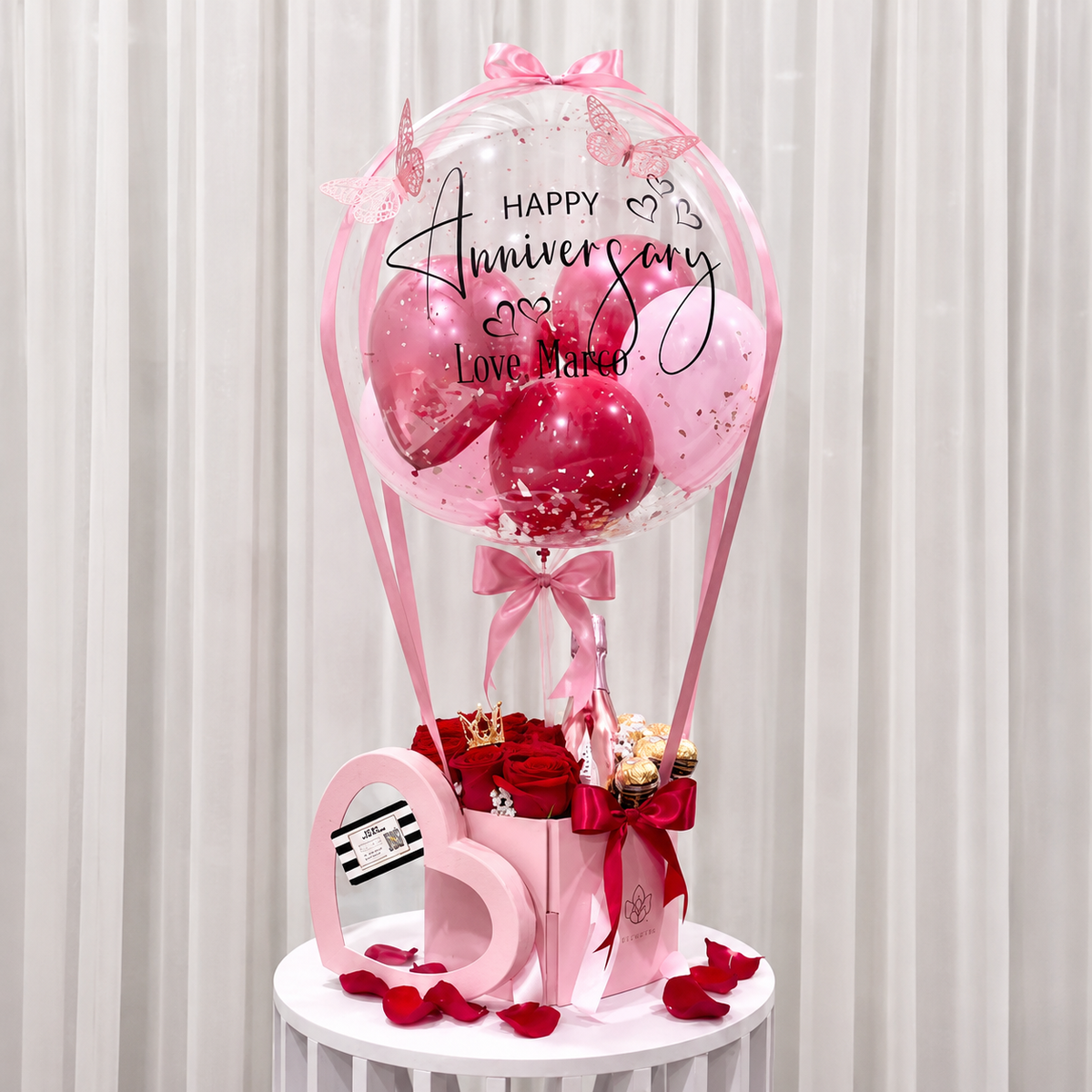 Romantic anniversary gift arrangement featuring a personalised clear balloon, pink and red balloons, red roses, chocolates, a pink bottle, and ribbon accents.