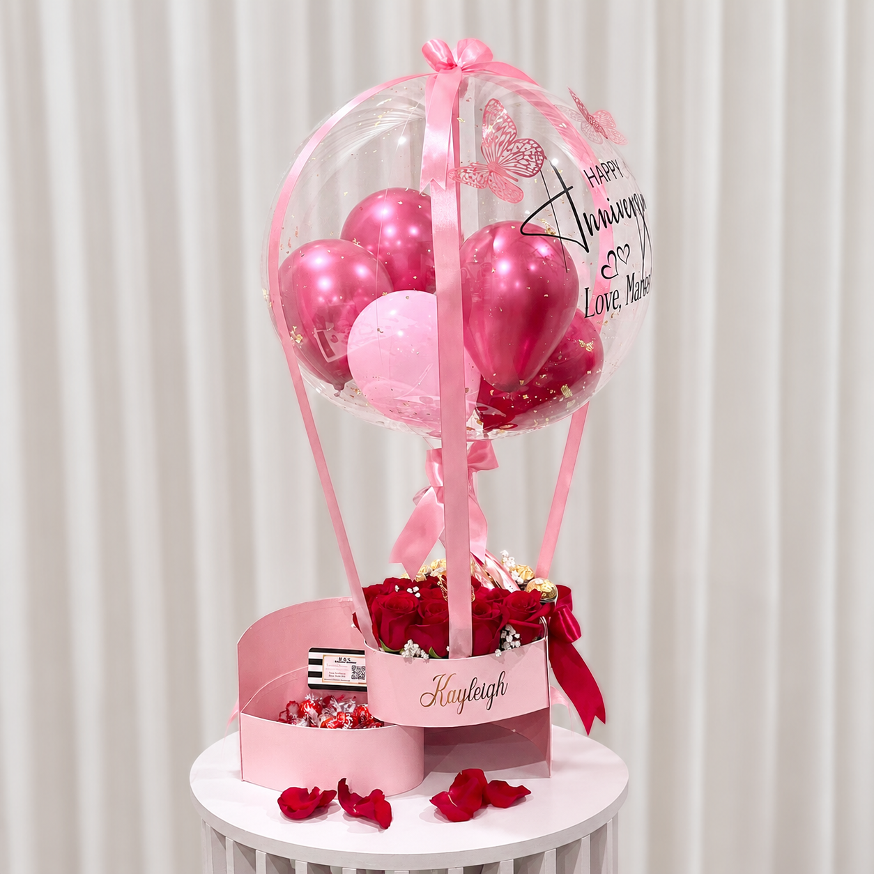Romantic anniversary gift arrangement featuring a personalised clear balloon, pink and red balloons, red roses, chocolates, a pink bottle, and ribbon accents.