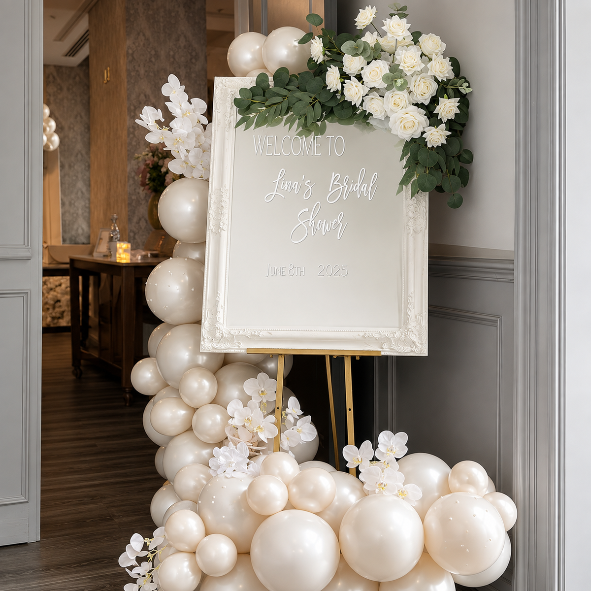 Lina’s bridal shower welcome display featuring a white floral sign, ivory balloon garland, white orchids, roses, eucalyptus greenery, and a gold easel.
