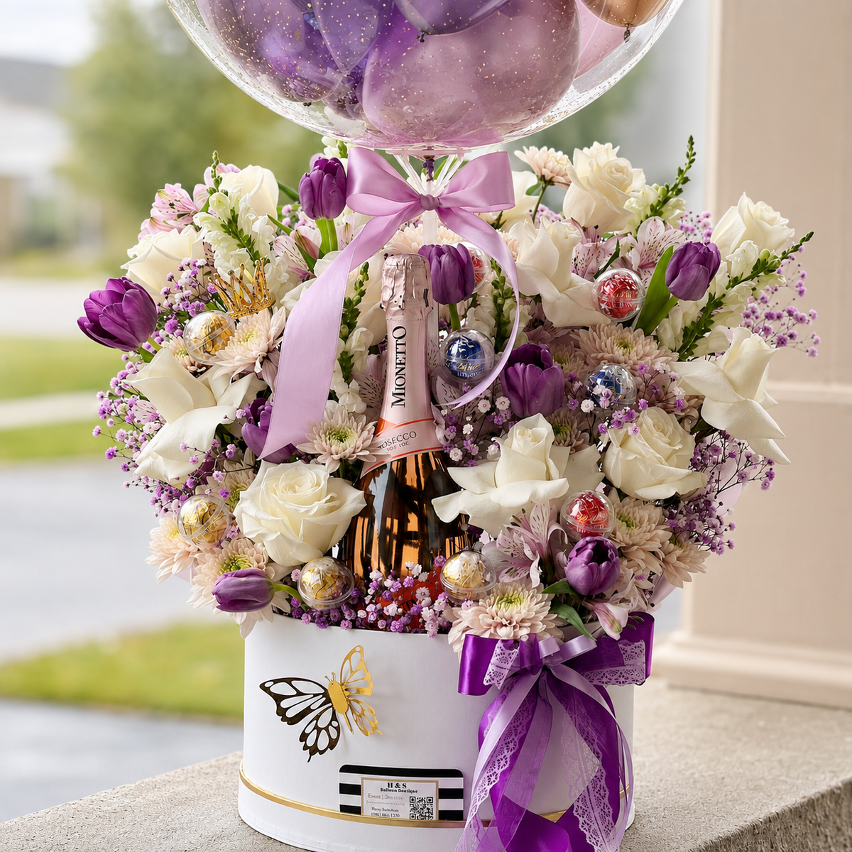 Luxury floral gift arrangement featuring purple tulips, white roses, decorative ornaments, a Prosecco bottle, and a clear balloon.