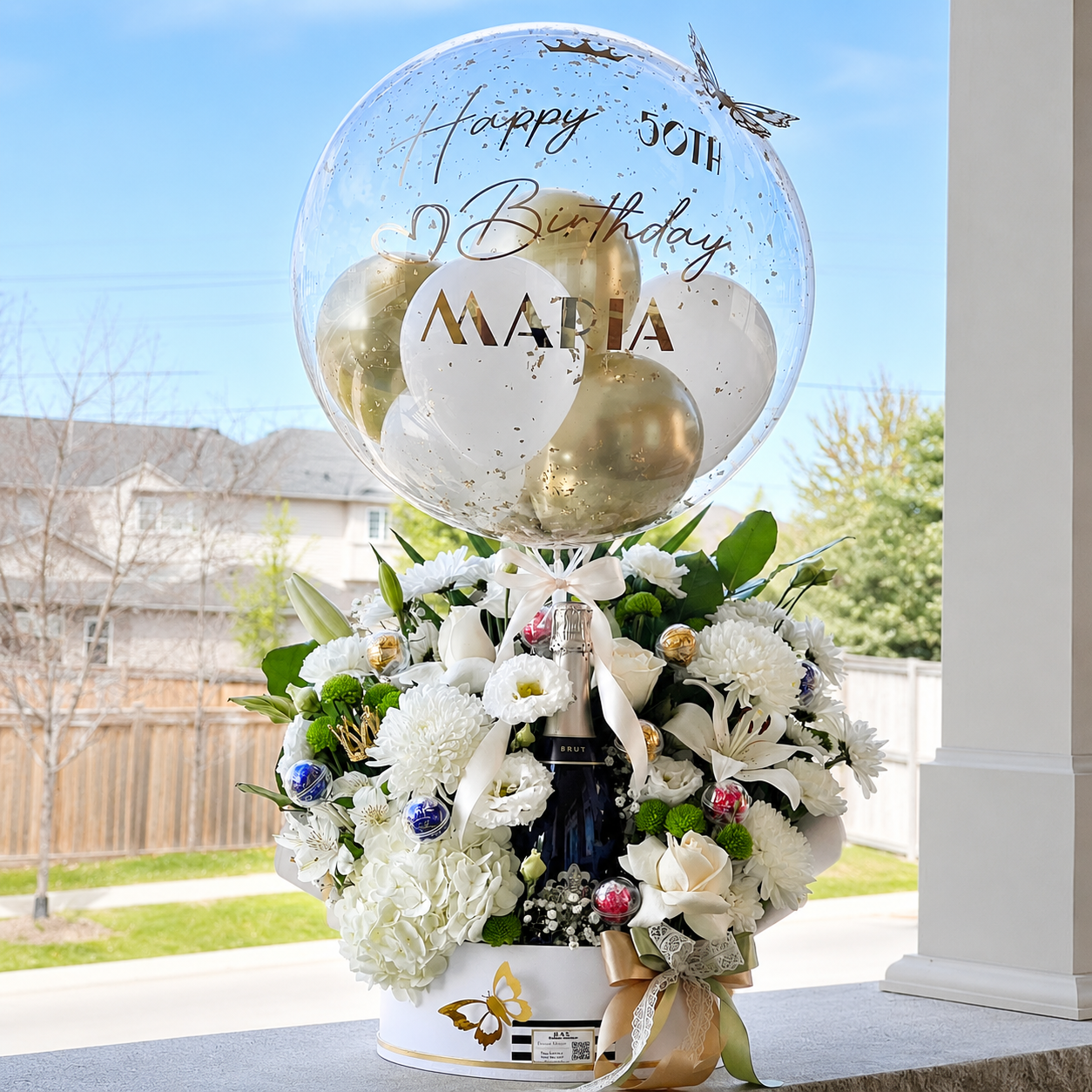 Luxury 50th birthday gift for Maria featuring a personalised clear balloon, white and gold balloons, fresh flowers, and a champagne bottle.