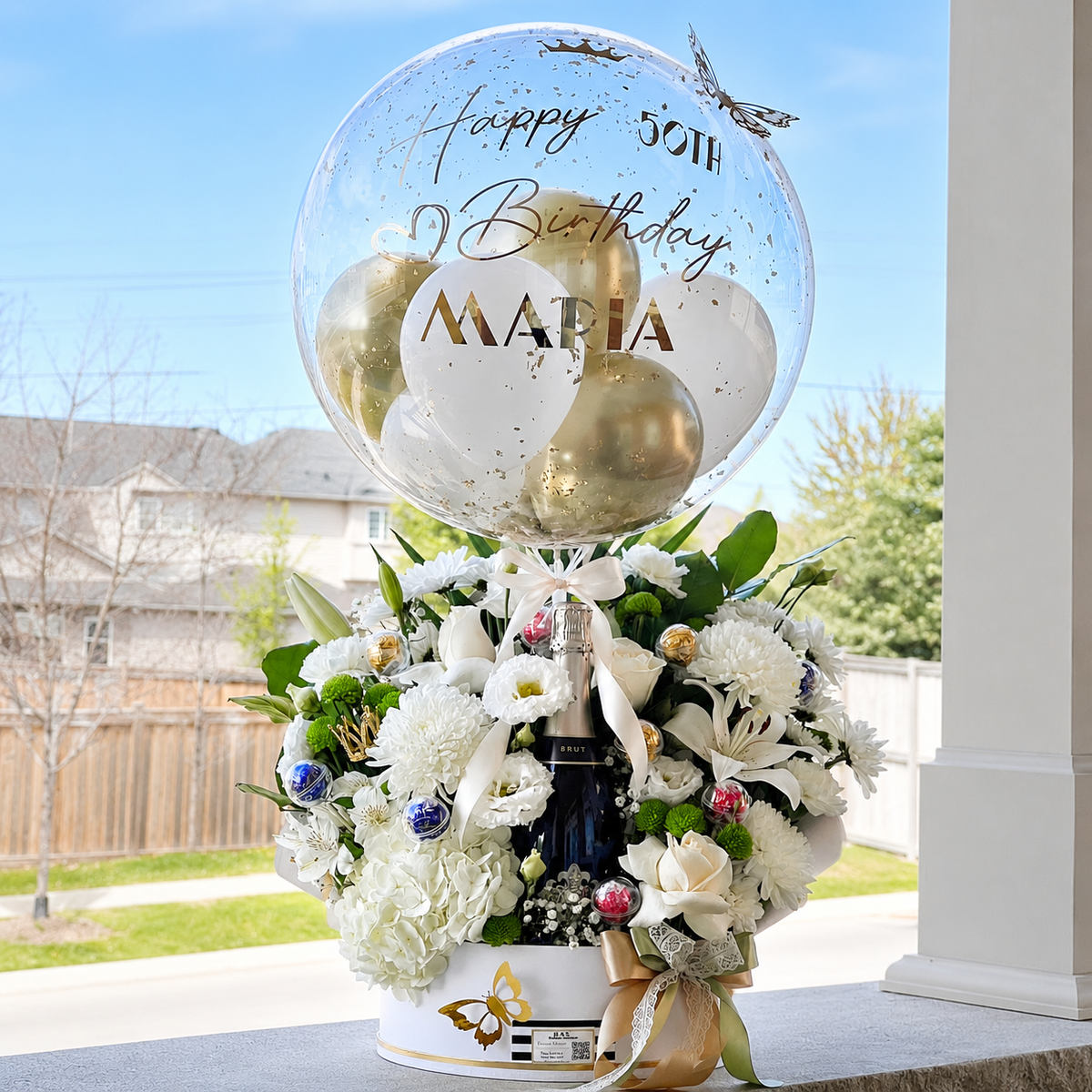Luxury 50th birthday gift for Maria featuring a personalised clear balloon, white and gold balloons, fresh flowers, and a champagne bottle.