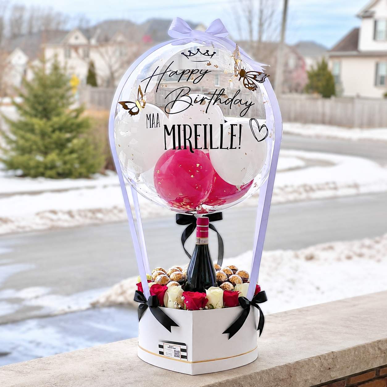Mireille’s birthday gift arrangement featuring a personalised clear balloon, pink and white balloons, roses, chocolates, a wine bottle, and black ribbon accents.