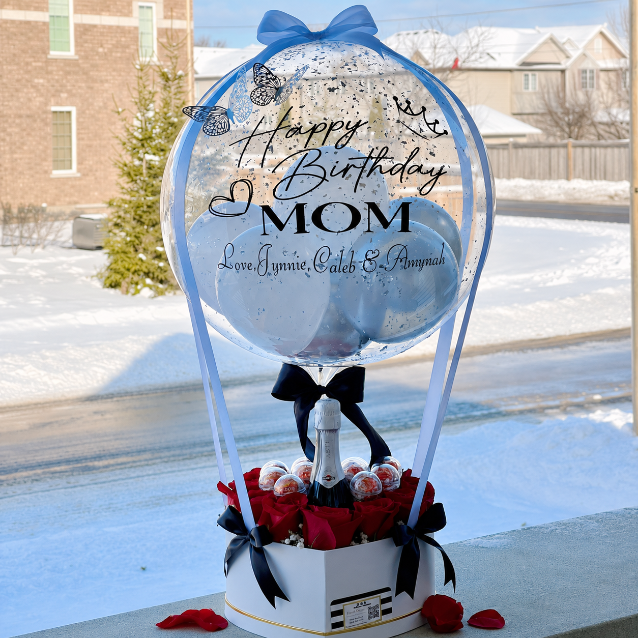 Birthday gift for Mom featuring a personalised blue balloon, red roses, chocolates, a champagne bottle, and black ribbon accents.