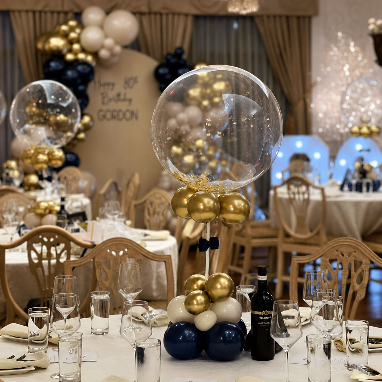 Elegant clear bubble balloon table centrepiece decorated with navy blue, gold, and cream balloons for an 80th birthday celebration.