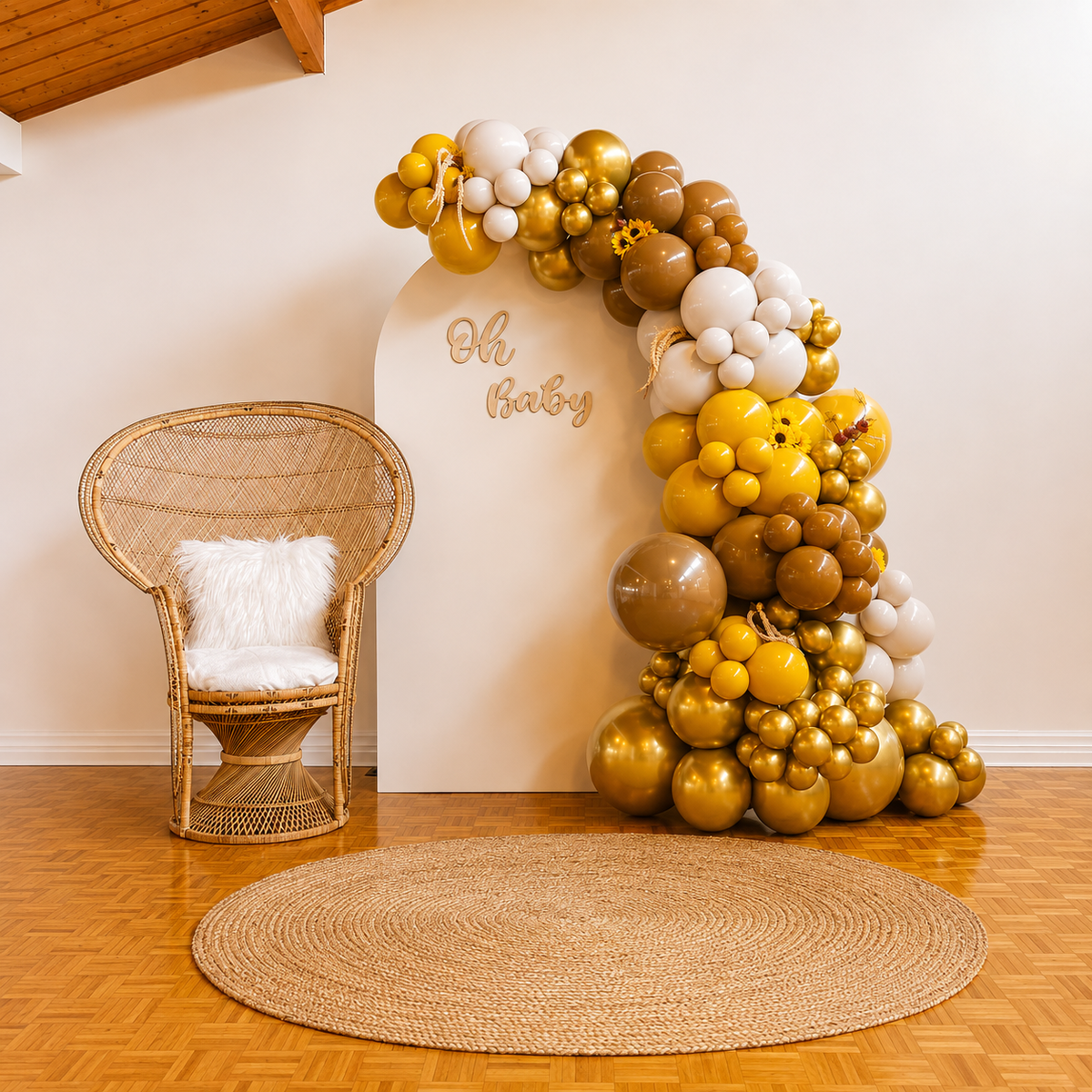 “Oh Baby” baby shower backdrop featuring mustard yellow, brown, cream, white, and gold balloon garlands with sunflower accents, a rattan peacock chair, and a woven rug.