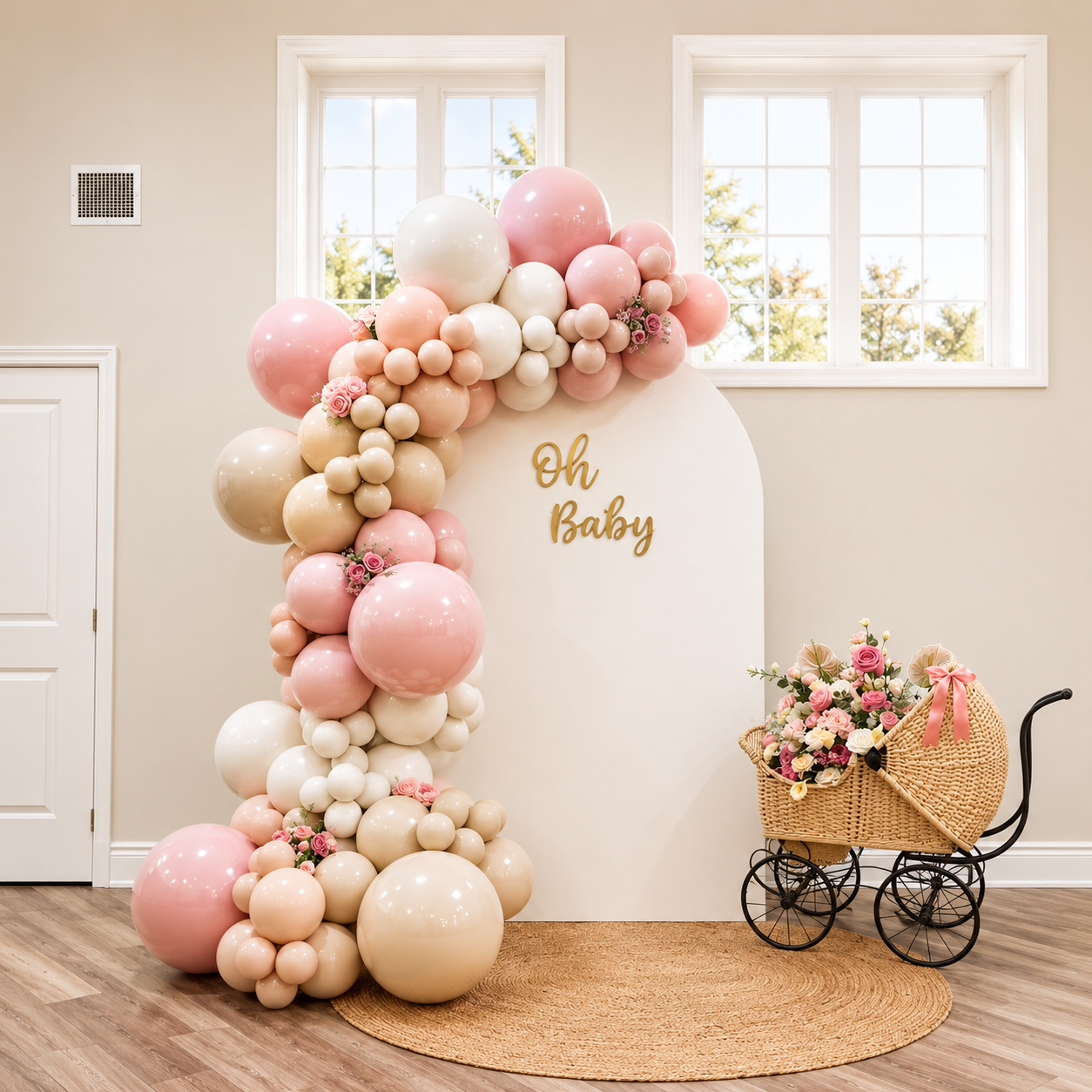 Elegant “Oh Baby” shower backdrop featuring pink, cream, and blush balloon garlands with floral accents, a white arched panel, woven rug, and vintage baby carriage filled with flowers.