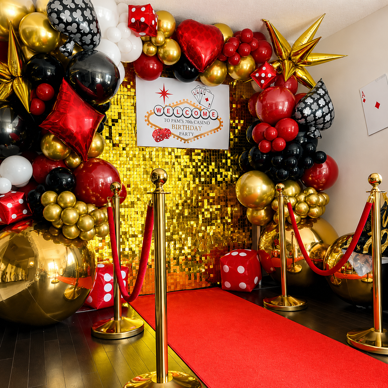 Pam’s 70th casino birthday backdrop featuring red, black, white, and gold balloons, playing card and dice decor, a gold shimmer wall, and a red carpet entrance.