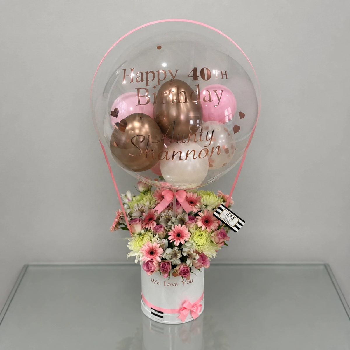 Personalized 40th birthday gift arrangement featuring a clear balloon with pink, white, and rose gold balloons, a mixed pink and white floral arrangement, heart accents, pink ribbon details, and a white “We Love You” gift box.