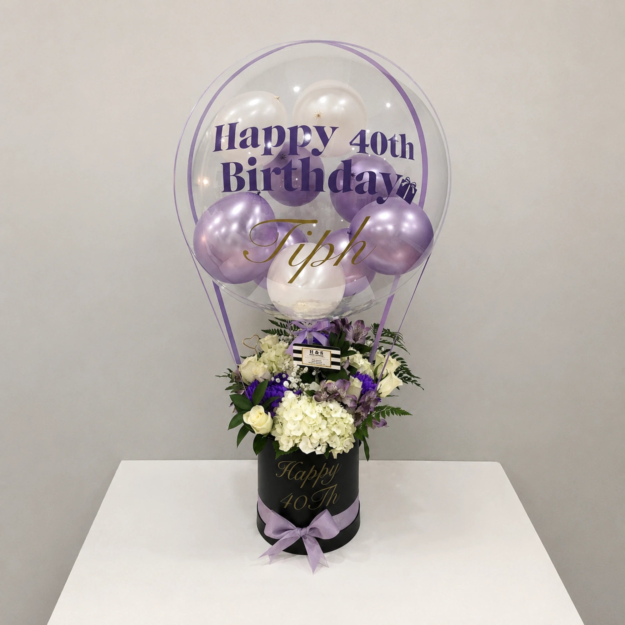 Personalized 40th birthday gift arrangement featuring a clear balloon with purple and white balloons, white hydrangeas, roses, purple flowers, greenery, a black “Happy 40th” gift box, and a lavender ribbon bow.