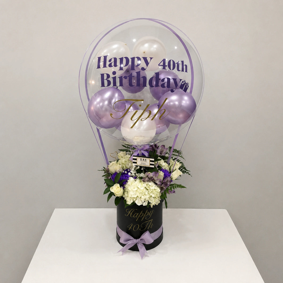 Personalized 40th birthday gift arrangement featuring a clear balloon with purple and white balloons, white hydrangeas, roses, purple flowers, greenery, a black “Happy 40th” gift box, and a lavender ribbon bow.