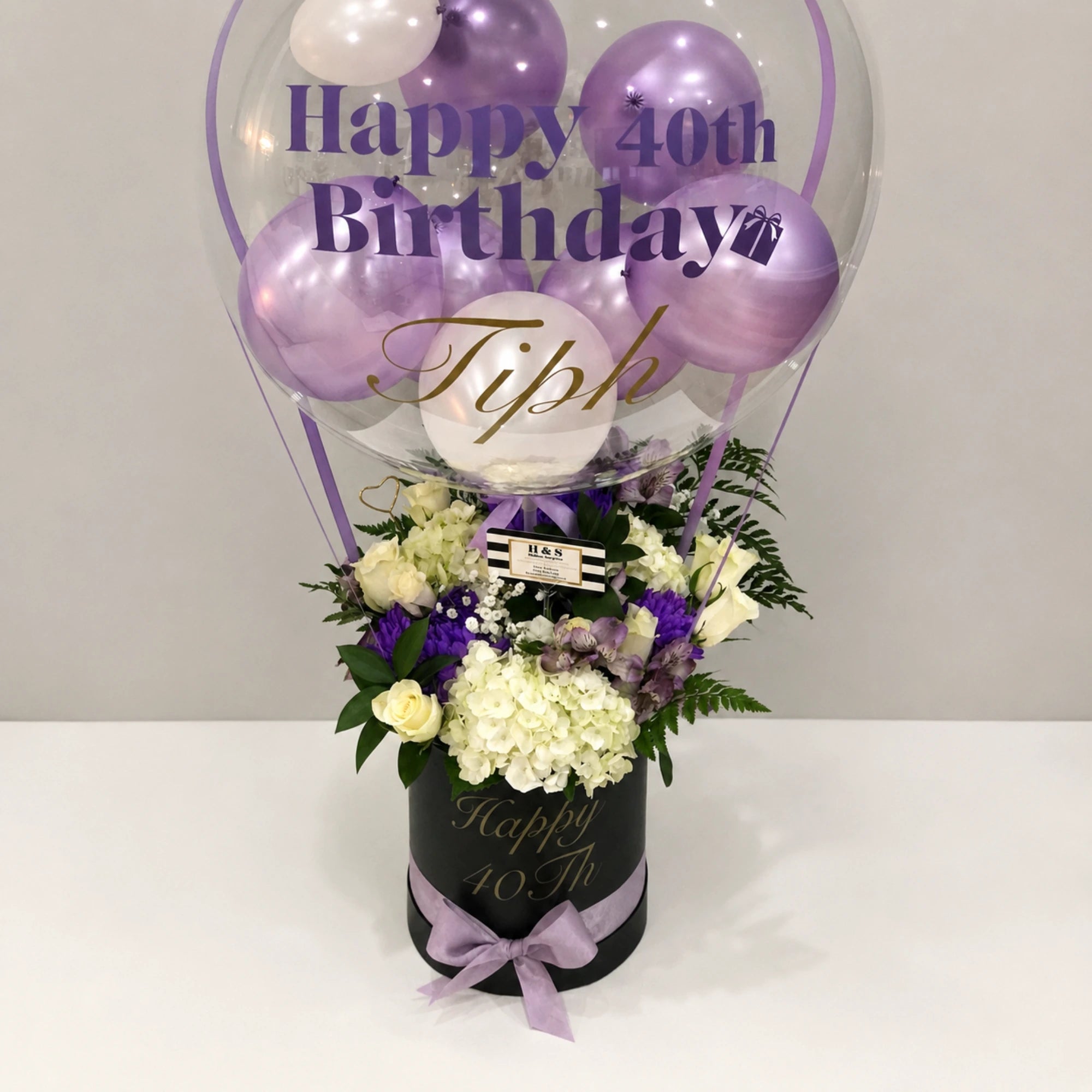 Personalized 40th birthday gift arrangement featuring a clear balloon with purple and white balloons, white hydrangeas, roses, purple flowers, greenery, a black “Happy 40th” gift box, and a lavender ribbon bow.