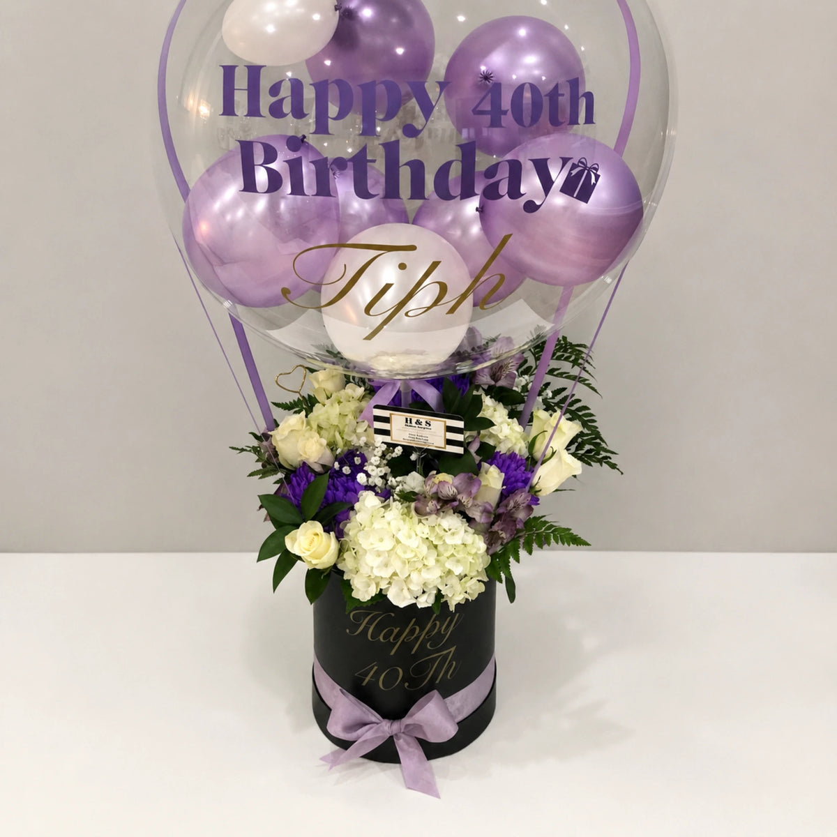 Close-up of a personalized 40th birthday gift arrangement featuring a clear balloon with purple and white balloons, white hydrangeas, cream roses, purple flowers, greenery, a black “Happy 40th” gift box, and a lavender bow.