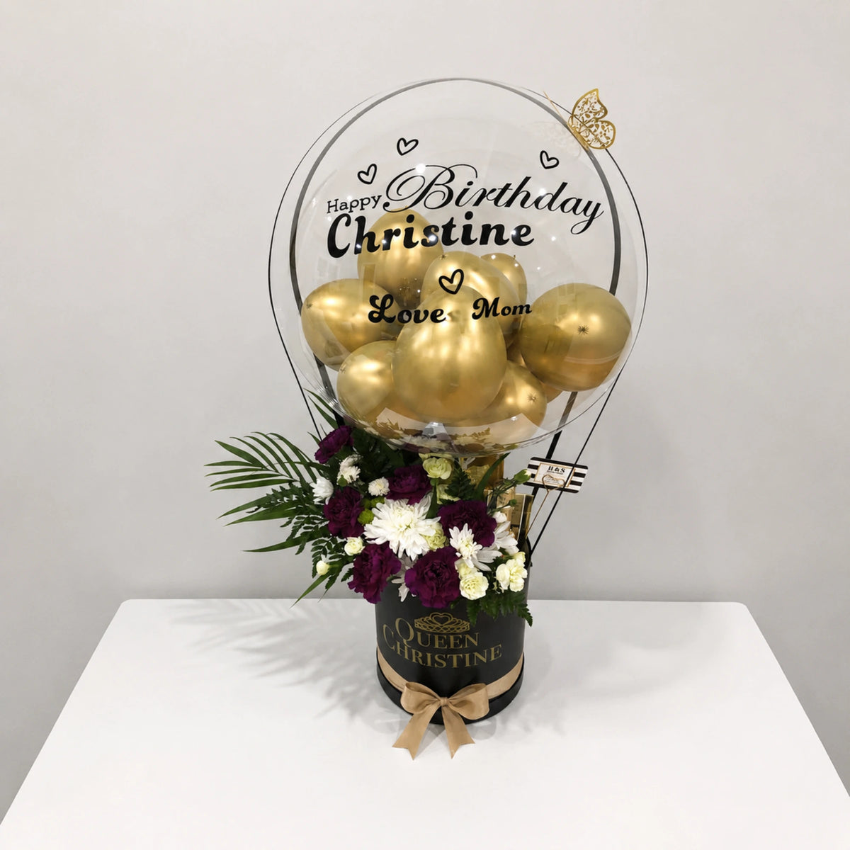 Personalized birthday gift arrangement for Christine featuring a clear balloon with gold balloons, purple and white flowers, greenery, gold butterfly accents, and a black “Queen Christine” gift box with a gold bow.