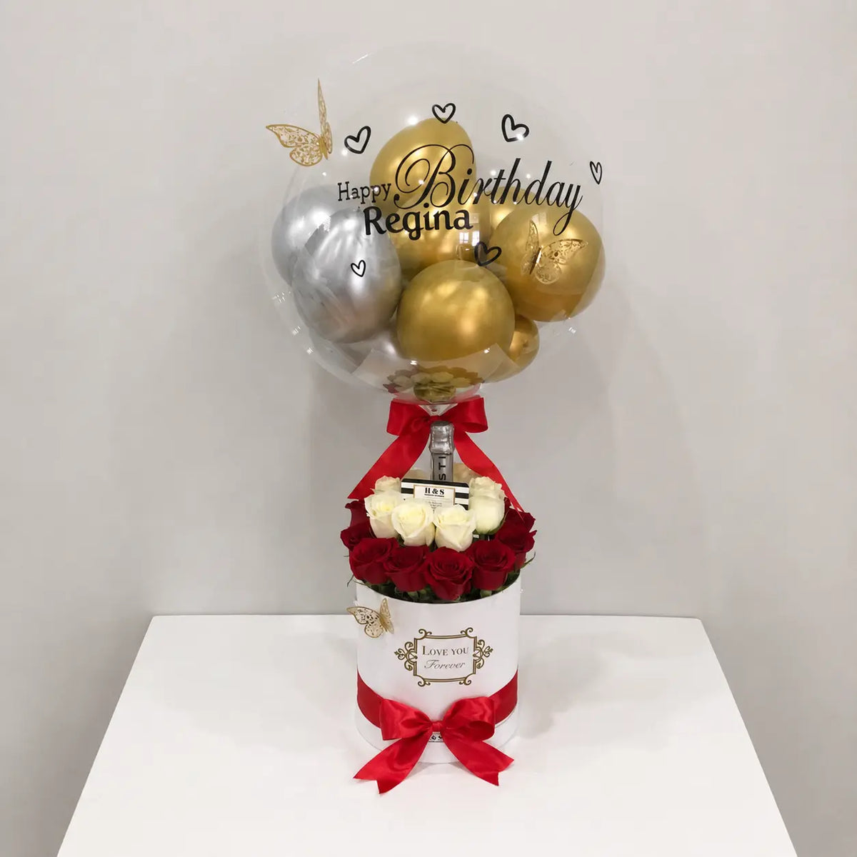 Personalized birthday gift arrangement for Regina featuring a clear balloon with gold and silver balloons, butterfly and heart accents, red and white roses, a bottle, white “Love You Forever” gift box, and red satin bows.