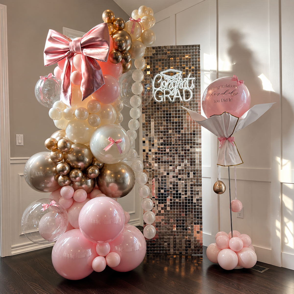 Graduation celebration backdrop featuring pink, white, and rose gold balloons, an oversized metallic bow, a rose gold shimmer wall, a glowing Congrats Grad sign, and a personalised balloon bouquet.