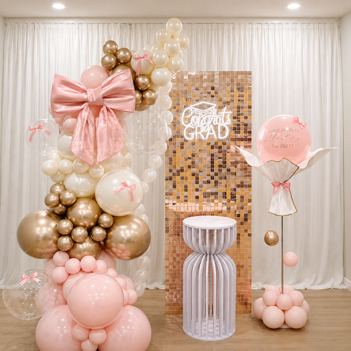 Graduation backdrop featuring blush pink, white, and rose gold balloons, an oversized satin bow, a rose gold shimmer wall, a glowing Congrats Grad sign, and a personalised pink balloon bouquet.