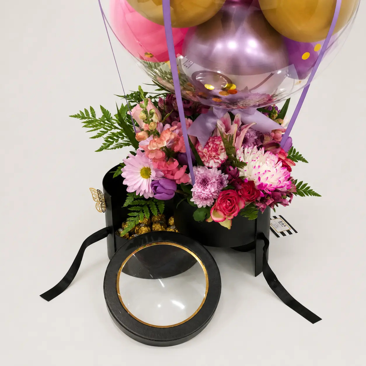 Close-up of a luxury birthday gift arrangement featuring pink and purple flowers, gold and purple balloons, greenery, gold-wrapped chocolates, butterfly accents, and a black gift box with ribbon details.