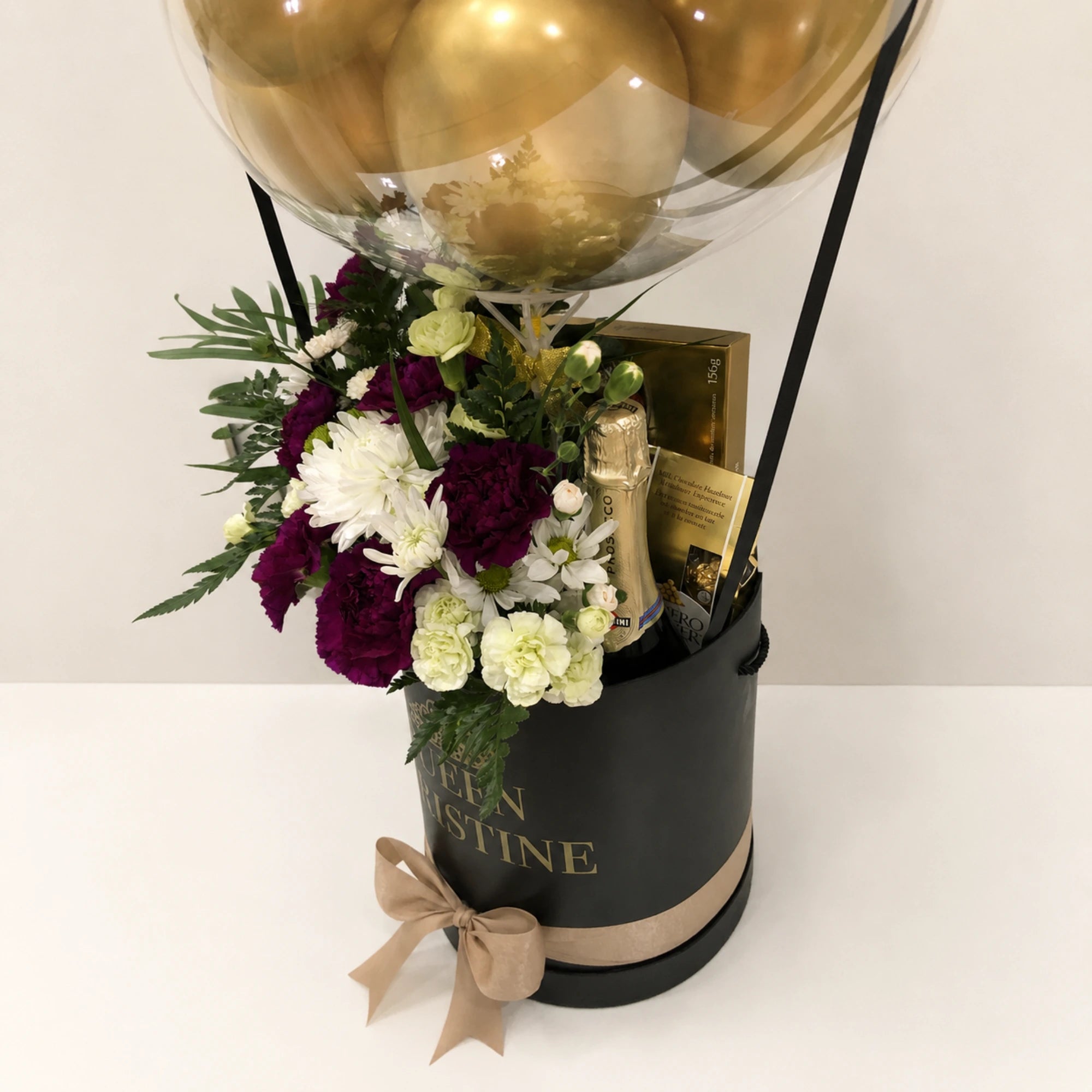 Personalized birthday gift arrangement for Christine featuring a clear balloon with gold balloons, purple and white flowers, greenery, gold butterfly accents, and a black “Queen Christine” gift box with a gold bow.