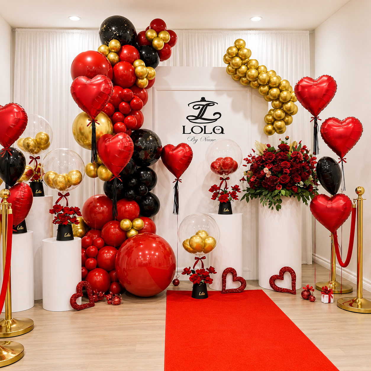 Elegant red, black, and gold event backdrop featuring heart-shaped balloons, balloon garlands, red roses, gold stanchions, clear bubble balloons, and a red carpet display.