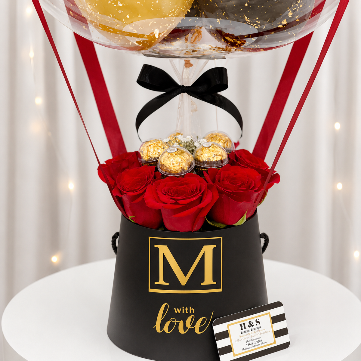 Luxury gift box featuring red roses, gold-wrapped chocolates, black and red ribbon accents, and a black box with gold “M With Love” detailing beneath a clear balloon.