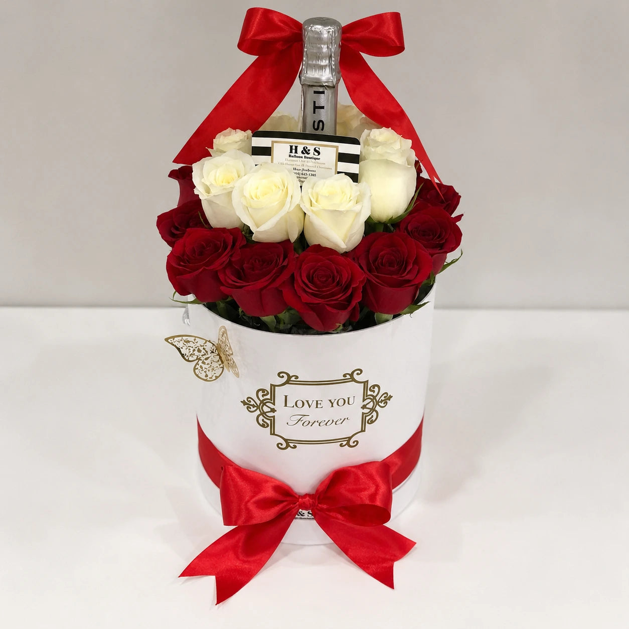 Personalized birthday gift arrangement for Regina featuring a clear balloon with gold and silver balloons, butterfly and heart accents, red and white roses, a bottle, white “Love You Forever” gift box, and red satin bows.