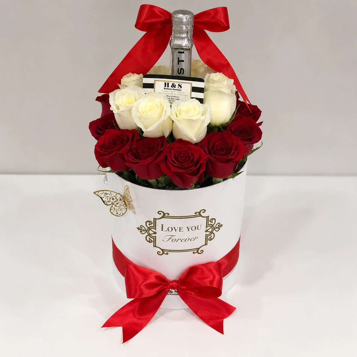 Romantic gift arrangement featuring red and white roses, a bottle, gold butterfly accent, red satin bows, and a white “Love You Forever” floral gift box.
